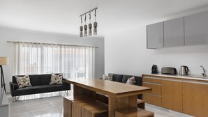 A modern living room with a wooden dining table, black sofa, and a kitchen area with grey cabinets and a coffee maker. - Lazy Days Adults Only Duna Parque Resorts & Hotels (Odemira)