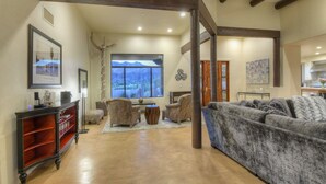 Un salon spacieux avec un grand canapé gris, une table basse en bois et deux fauteuils à motifs. Une console en bois, dotée d’étagères, est adossée au mur, et une grande fenêtre offre une vue sur les montagnes.