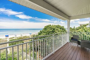 Um deck de madeira com guarda-corpos brancos, com vista para uma praia com guarda-sóis e o oceano.
