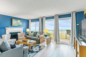 A living room with a fireplace, a sofa, a coffee table, and a TV. There is a balcony with a view of the beach. - Beach Cottages I 207 - GULF VIEW Condo!! (Indian Shores)