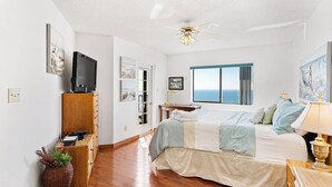A bedroom with a bed, a television, a ceiling fan, and a window with a view of the ocean.