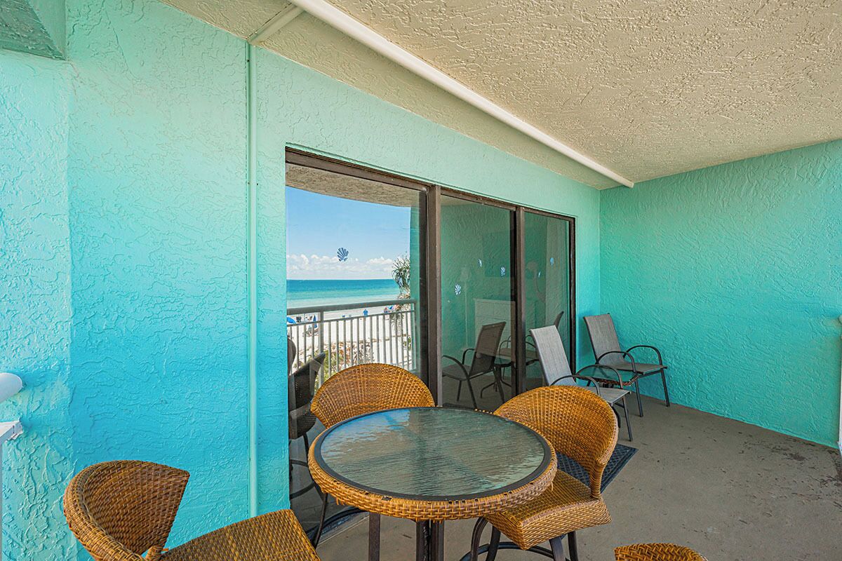 A balcony with wicker chairs and a glass table, overlooking a beach with a clear view of the ocean and palm trees.
