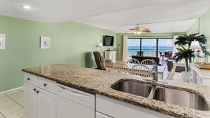 A kitchen with granite countertops, a built-in dishwasher, and a large sink. There is a dining area with a table and chairs, and a view of the ocean through large windows.