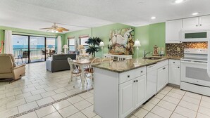 A spacious living area with a dining table, chairs, and a sofa. A kitchen with white cabinets and a large island is visible.