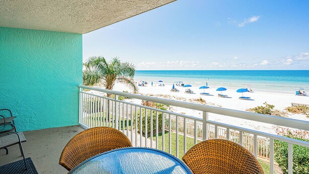 A balcony with wicker chairs and a table, overlooking a beach with umbrellas and people.