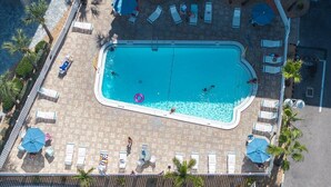 Eine Vogelperspektive zeigt einen rechteckigen Pool mit Liegestühlen und Sonnenschirmen, umgeben von Palmen und einer gefliesten Terrasse.