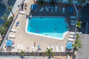 Ein rechteckiger Swimmingpool, umgeben von Liegestühlen und Sonnenschirmen, mit Palmen und einem gepflasterten Weg.