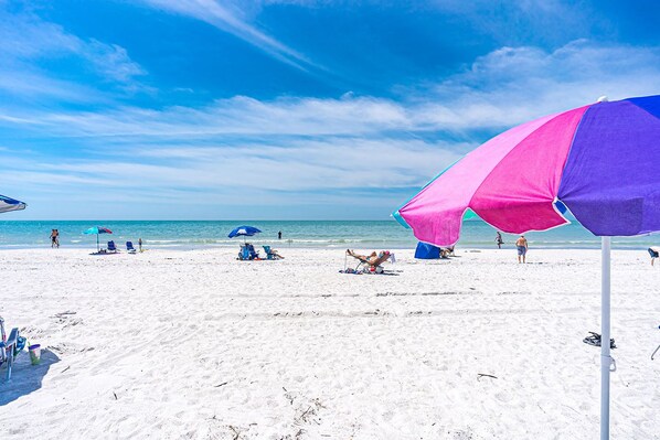 A beach with white sand and clear blue water. There are several beach umbrellas and lounge chairs.
