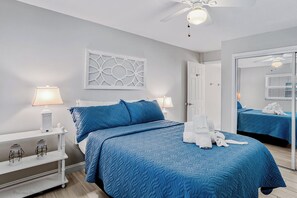 Quarto de hotel com uma cama, duas luminárias de cabeceira, uma mesa de cabeceira branca, um espelho e um ventilador de teto.
