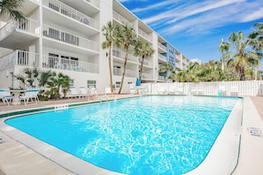 A swimming pool surrounded by a white building with balconies, palm trees, and lounge chairs.