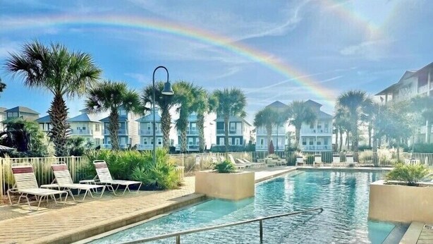 A pool area with lounge chairs, palm trees, and a building in the background. - Fall Rates! | Pool | Foosball | Dog Friendly (Mexico Beach)