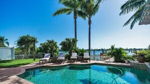 A swimming pool with lounge chairs and palm trees, surrounded by a fence and a view of the water. - Ohana House 333 - Intercoastal Views with a Pool (Indian Rocks Beach)
