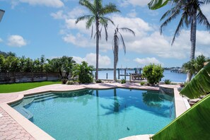 A swimming pool with a curved edge, surrounded by palm trees and a view of the water. - Ohana House 333 - Intercoastal Views with a Pool (Indian Rocks Beach)