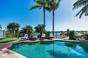 A swimming pool with lounge chairs and palm trees, surrounded by a fence and a view of the water.
