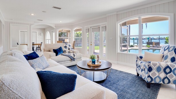 A spacious living room with a large window overlooking a pool, a white and blue patterned armchair, and a beige sofa with blue cushions. - Ohana House 333 - Intercoastal Views with a Pool (Indian Rocks Beach)