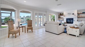 A spacious living room with a white sofa, two wicker chairs, a small table, and a fireplace. Large windows offer views of a pool and greenery. - Ohana House 333 - Intercoastal Views with a Pool (Indian Rocks Beach)