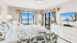 A hotel room with a bed, a TV, a balcony with a view of the ocean, and a ceiling fan. - Gulf-Front 3BR | Pool & Sunsets | Reef Club 310 (Indian Rocks Beach)