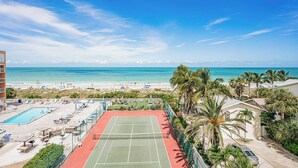 A tennis court with a pool, beach view, and palm trees.