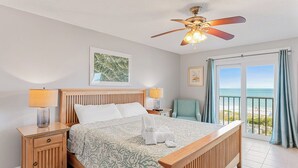 A bedroom with a bed, two bedside tables, a ceiling fan, a window with curtains, and a view of the beach.