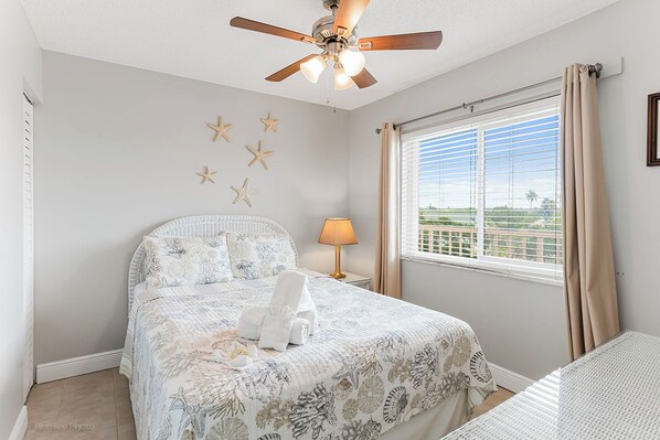 Un dormitorio con una cama, ventilador de techo, mesita de noche con lámpara, decoración de paredes con motivos de estrellas de mar y una ventana con cortinas.