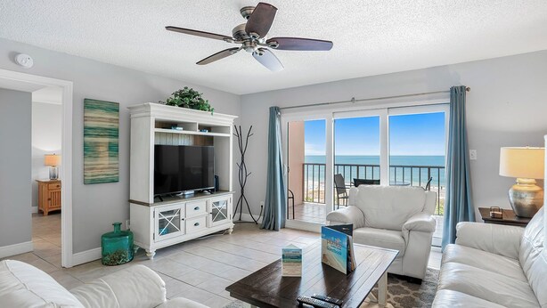 A spacious living room with a television, white furniture, a ceiling fan, and a balcony with a view of the beach.