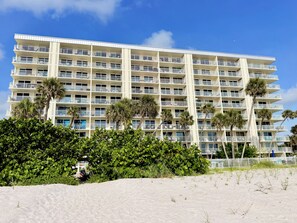 A multi-story hotel with balconies, palm trees, and a sandy foreground.