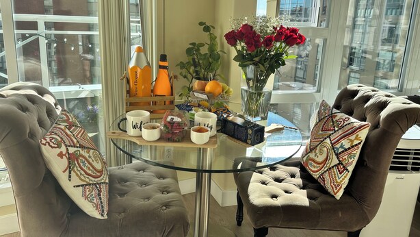 Un salon moderne avec une table en verre, deux fauteuils marron, une machine à café, un vase de roses rouges et une bouteille de jus d’orange.