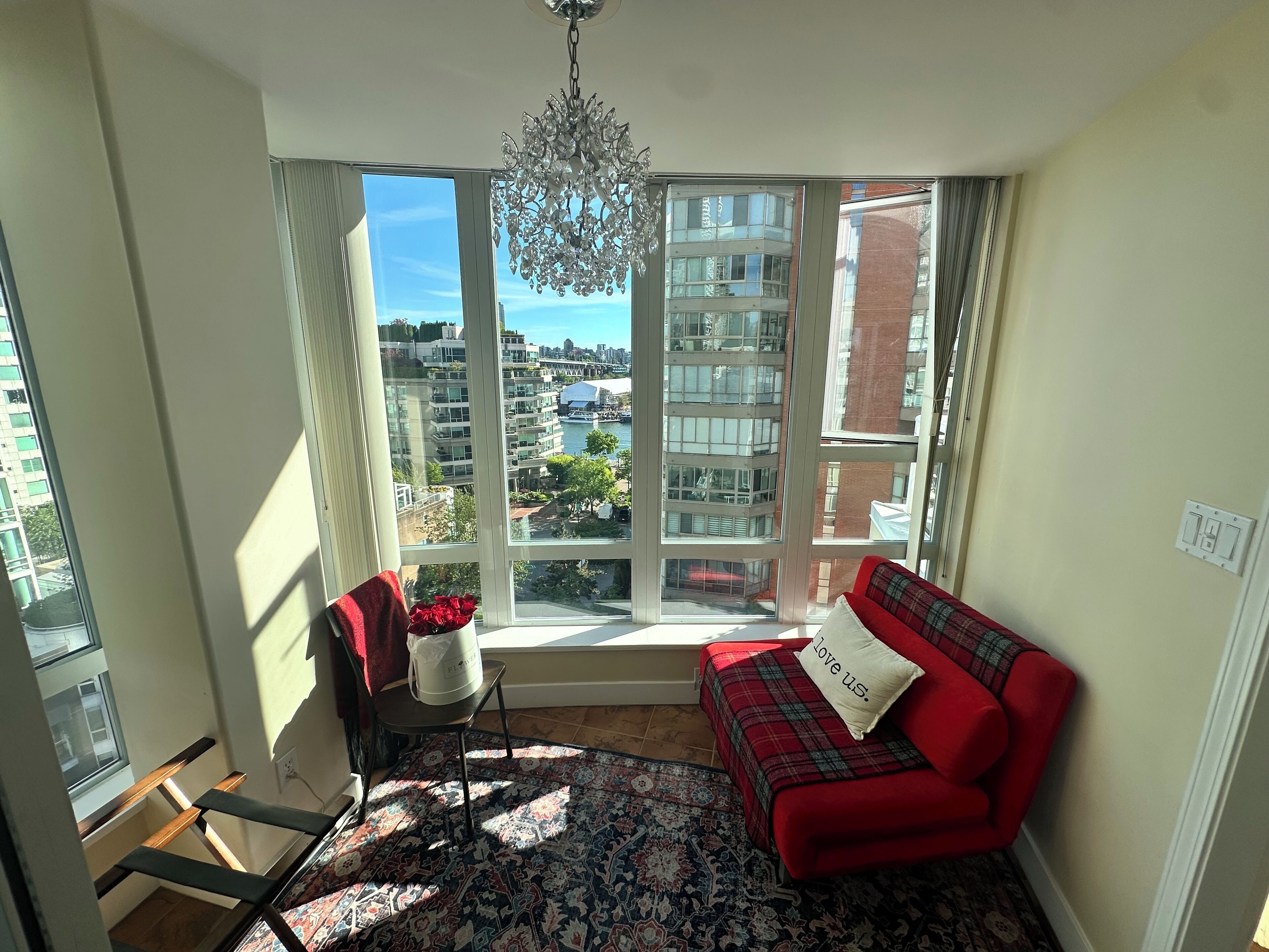 Quarto com sofá vermelho, tapete estampado, uma cadeira, uma mesinha com um vaso de flores e uma janela grande com vista para edifícios e um corpo d'água.