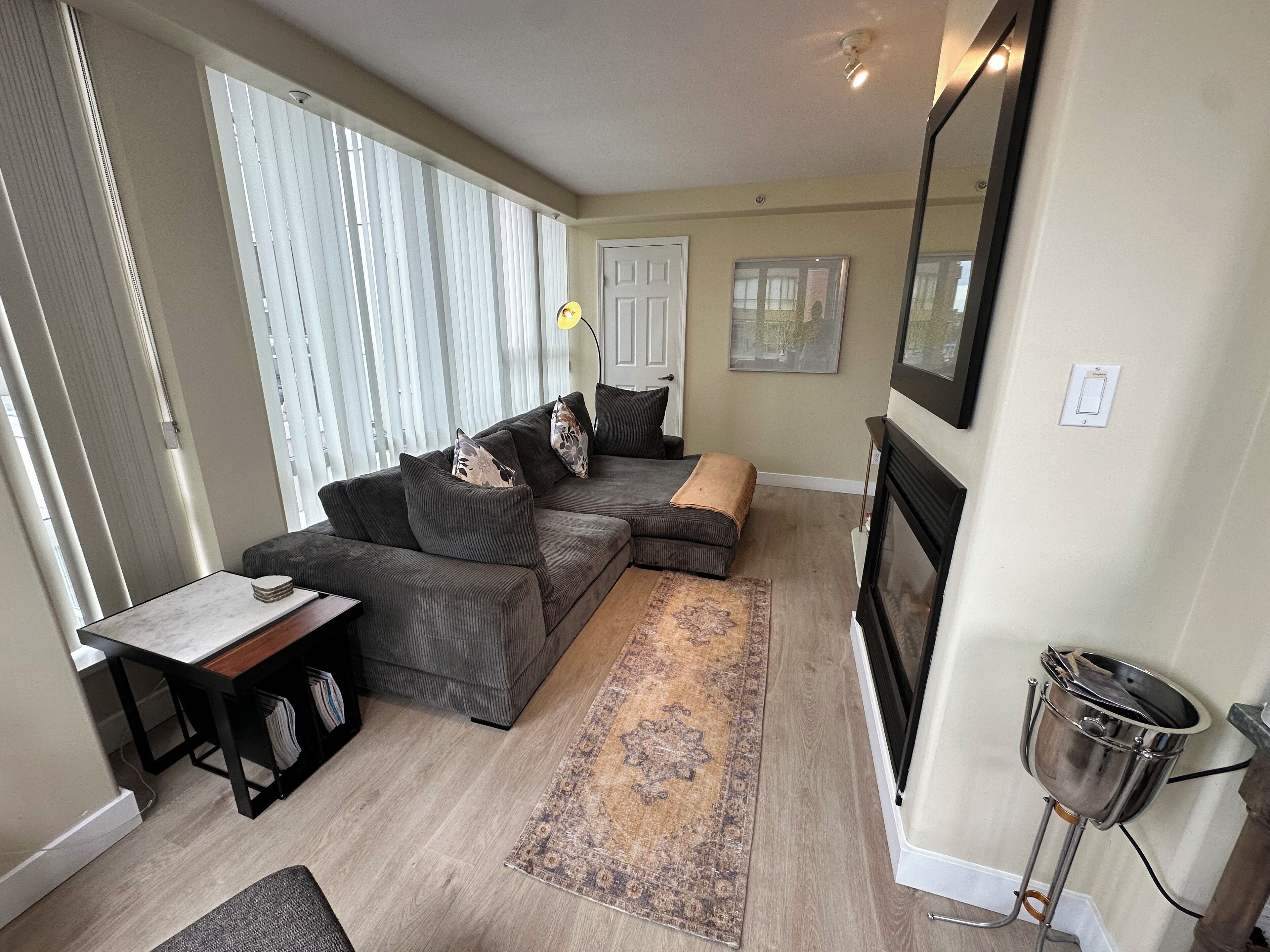 A living room with a sectional sofa, a small table, a rug, a fireplace, and a window with blinds.