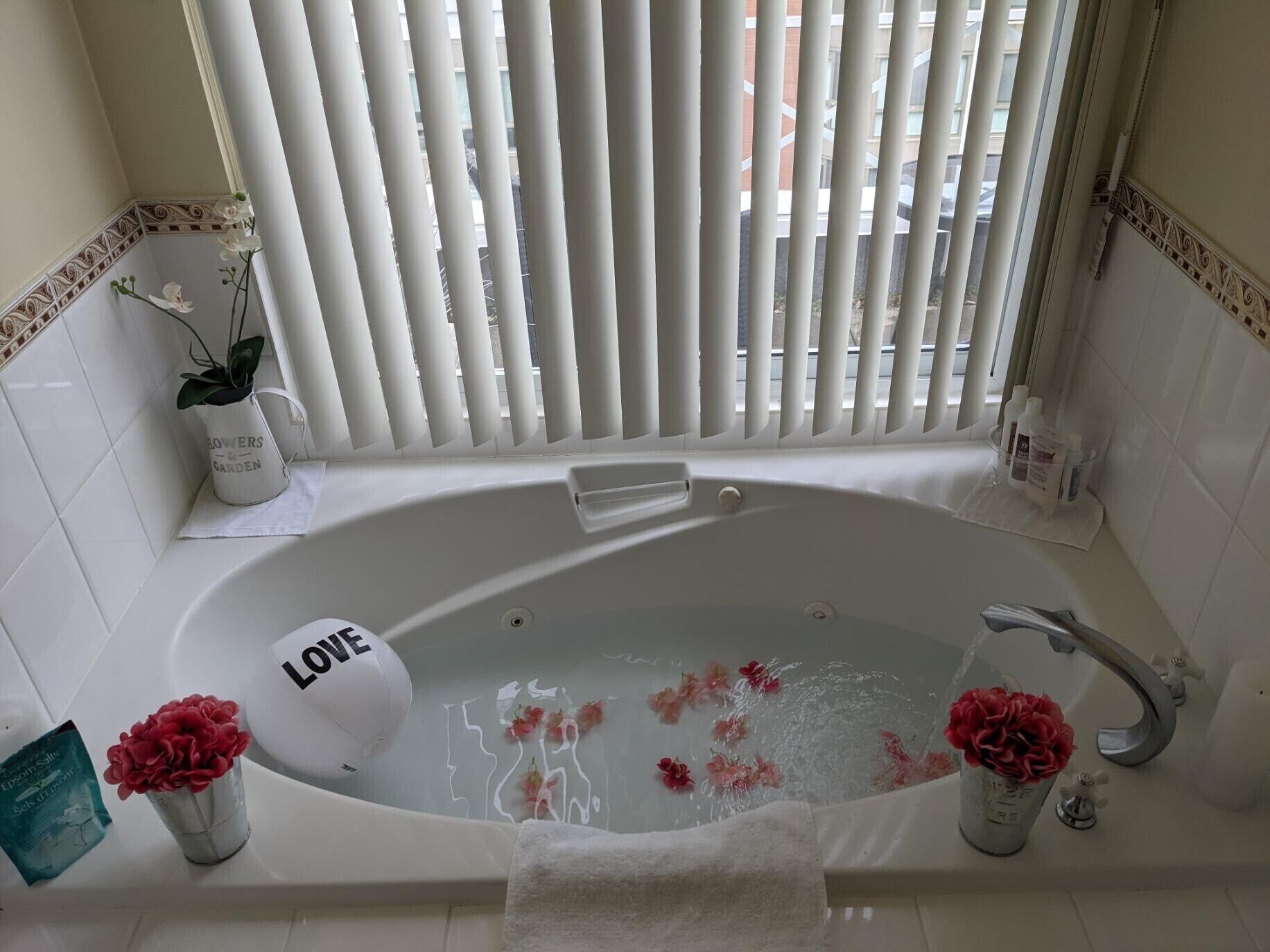 Une salle de bain avec une baignoire, une douche et une fenêtre avec des stores. Il y a deux vases à fleurs sur le sol.