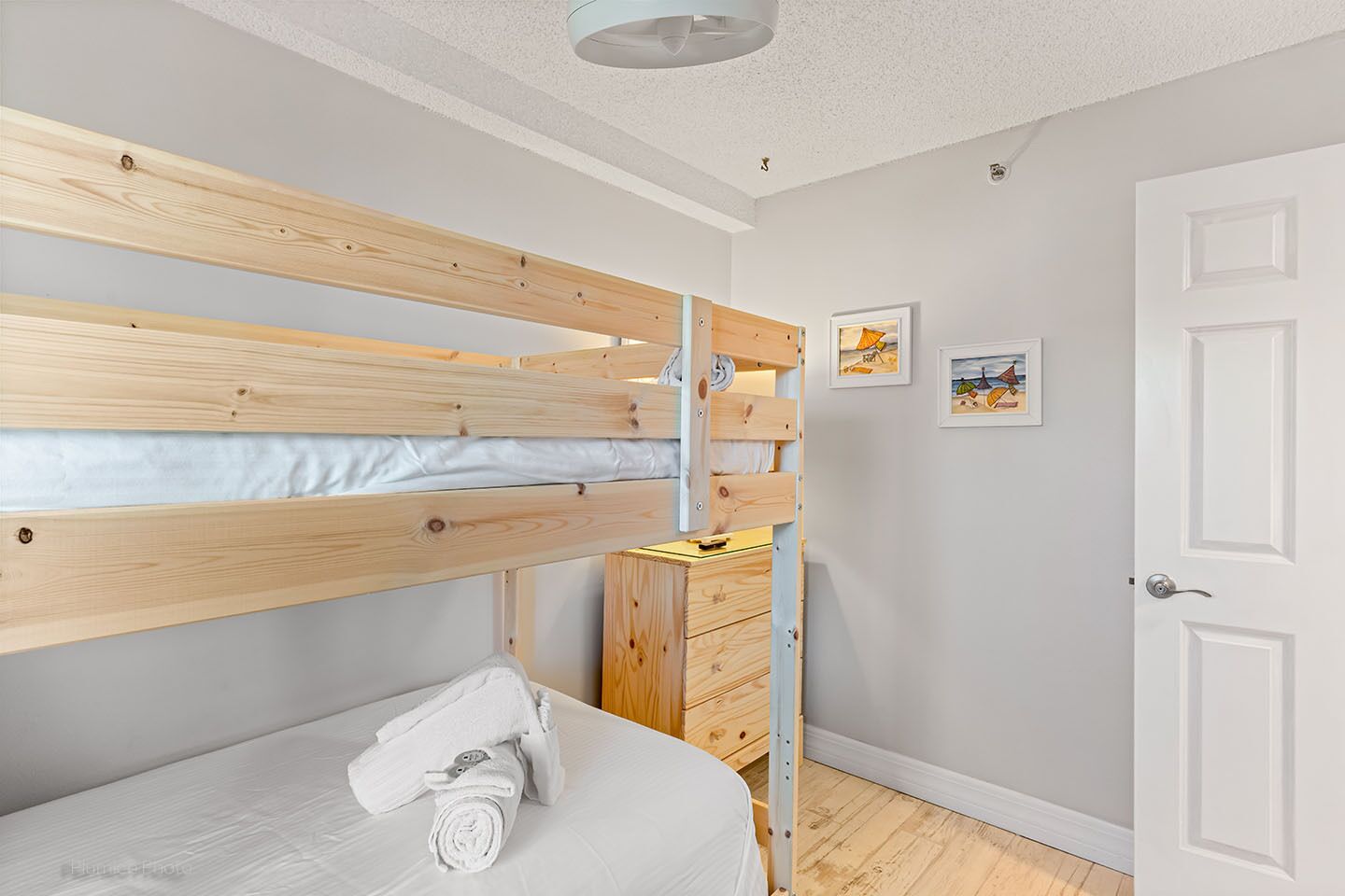 Un lit superposé avec une structure en bois, un matelas blanc et une serviette enroulée sur le lit du bas. On y trouve une commode en bois avec des tiroirs, une porte blanche et deux tableaux encadrés accrochés au mur.