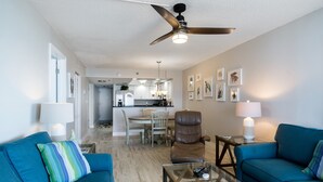Un salon avec deux canapés bleus, une table basse en verre, un ventilateur de plafond et, au fond, une cuisine.