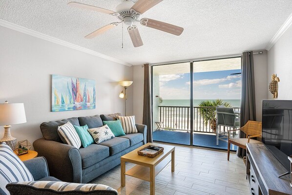 A living room with a sofa, a coffee table, a TV, and a balcony with a view of the ocean.