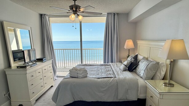 Ein Schlafzimmer mit einem Bett, Nachttischen, einem Schminktisch und einem Deckenventilator, mit Blick auf das Meer.