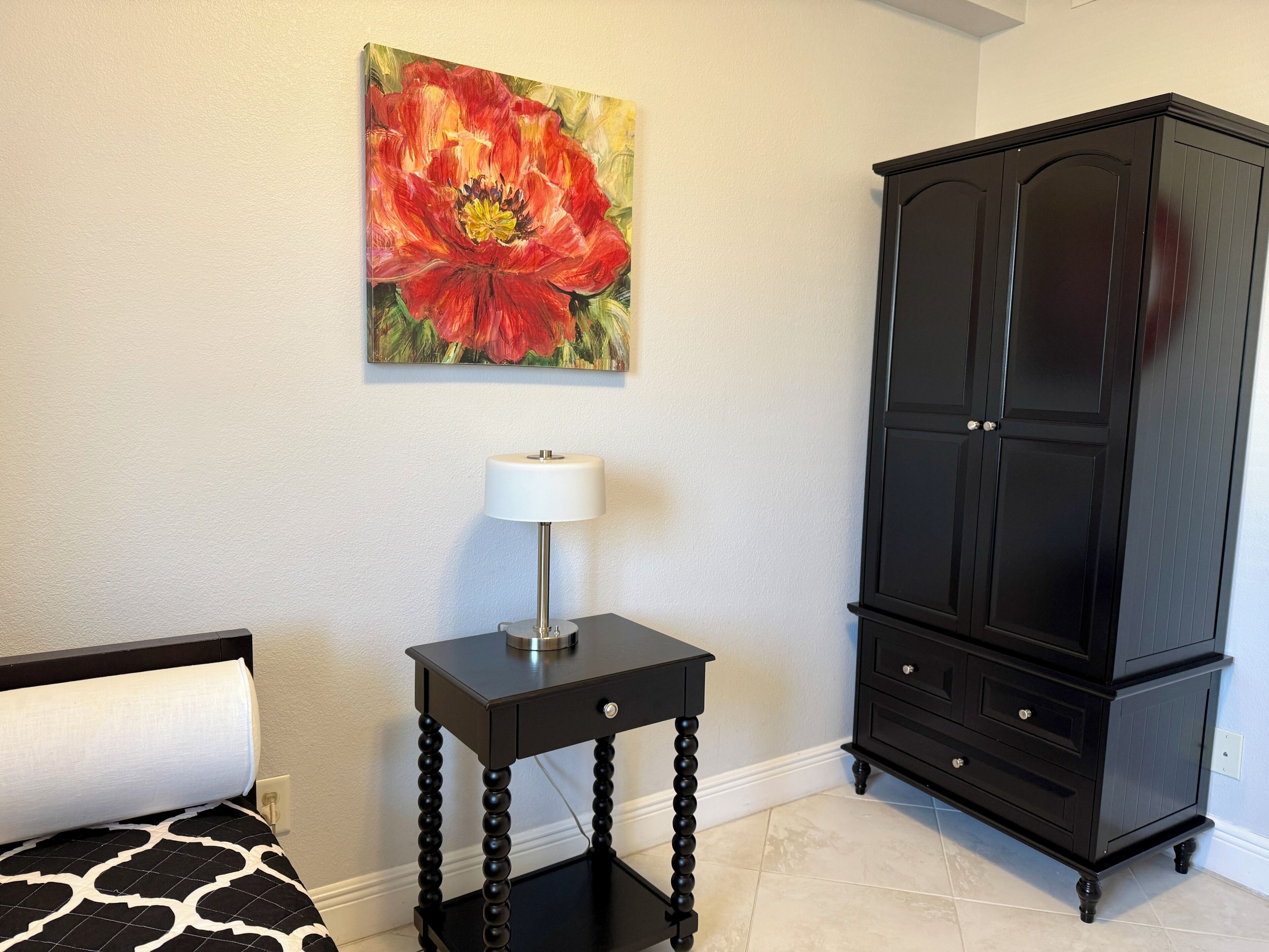 A bedroom with a bed, a small table with a lamp, a black cabinet, and a painting on the wall.
