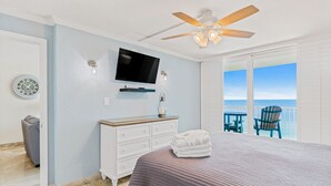 A bedroom with a bed, a dresser, a television, a ceiling fan, a clock, and a view of the ocean.