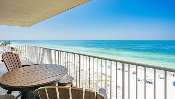 A balcony with a wooden table and chairs, overlooking a beach with clear blue water and a white railing.