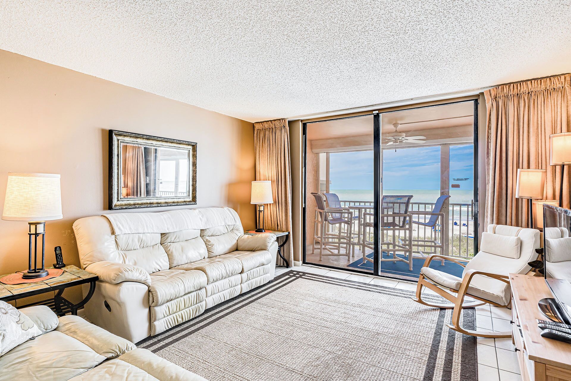A hotel room with a large sliding glass door leading to a balcony with beach views. There is a sofa, a small table with a lamp, and a chair.