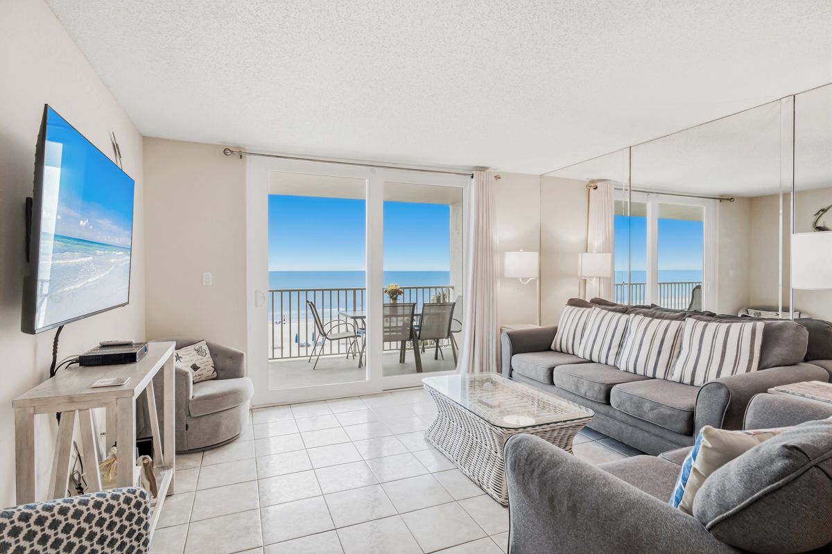 A modern living room with a sofa, a coffee table, and a TV. There is a balcony with a view of the sea.