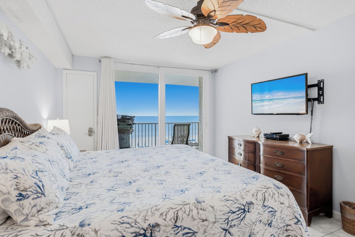 A bedroom with a large bed, a ceiling fan, a television, and a view of the ocean through a sliding door.