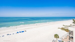 Ein Strand mit weißem Sand, klarem, blauem Wasser und einigen Spaziergängern. Entlang der Küste stehen blaue Sonnenschirme und Liegestühle.