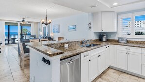 A modern kitchen with white cabinets, a granite countertop, and a dishwasher. There is a large window with a view of buildings and a balcony with outdoor furniture.