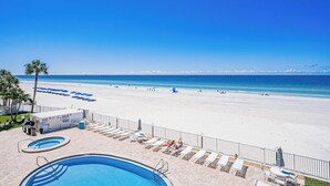 A pool area with lounge chairs and umbrellas, a beach with people and umbrellas, and a clear blue sky.