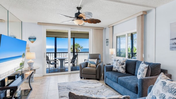 A spacious living room with a large sofa, a coffee table, and a TV. There is a balcony with a view of the ocean.