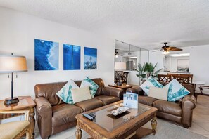 A living room with a brown leather sofa, a glass coffee table, and a wooden side table. There are three wall-mounted paintings, a ceiling fan, and a dining area in the background. - Sand Castle III 403 - GULF FRONT Condo! (Indian Shores)