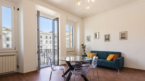A spacious living area with a glass table, clear chairs, a blue sofa, and a view of buildings through large windows. - Santa Croce View 1 by Mmega (Firenze)