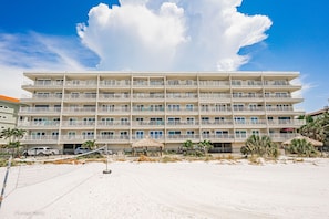 A multi-story hotel with balconies, located on a sandy beach with palm trees and a thatched-roof structure.