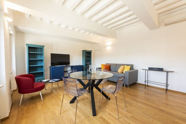 A modern living room with a glass-top dining table, red and blue chairs, a grey sofa, and a flat-screen TV.