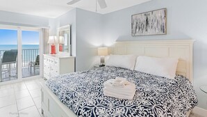 A bedroom with a bed, a nightstand, a dresser, a ceiling fan, and a view of the ocean through a balcony.