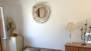 A bedroom with a bed, a wooden dresser, a wall-mounted mirror, a lamp, and a decorative wall piece. - Adorable T2 apartment (45m2) in a residence with panoramic sea view (Jard-sur-Mer)